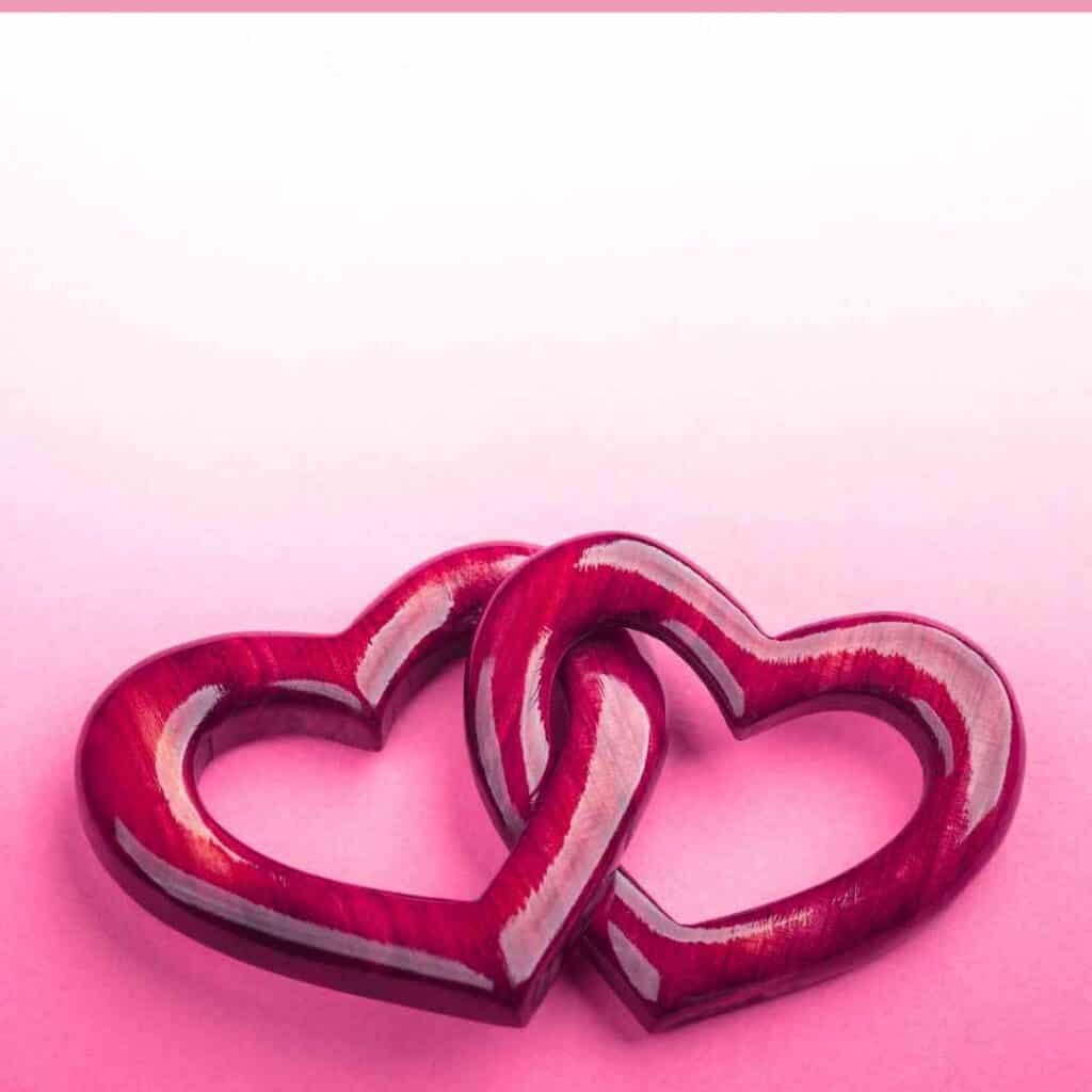 Photograph of 2 wooden hearts that are a deep red color. The hearts are intertwined and are on a background that is pink on the bottom and fades to white at the top. There is pink broborder at the top of the picture. Used to illustrate family love for the post prayers for my granddaughter.