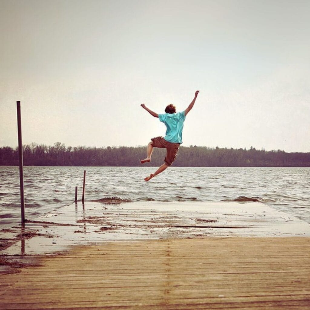 Photo of a teen age boy jumping off a pier into a lake. He has his arms and legs spread in a joyful manner. Used to illustrate the post Praying for my Grandson, to show an activity a boy might like to do.