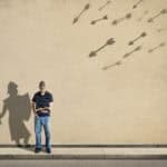 Featured image for the Armor of God post. The picture shows a young man dressed in blue jeans, a t-shirt, and a baseball hat. He is standing up and is reading a Bible in his hand. The shadow behind shows a warrior with a sword and shield. In the top right of the picture, arrows of different sizes are flying towards the young man.
