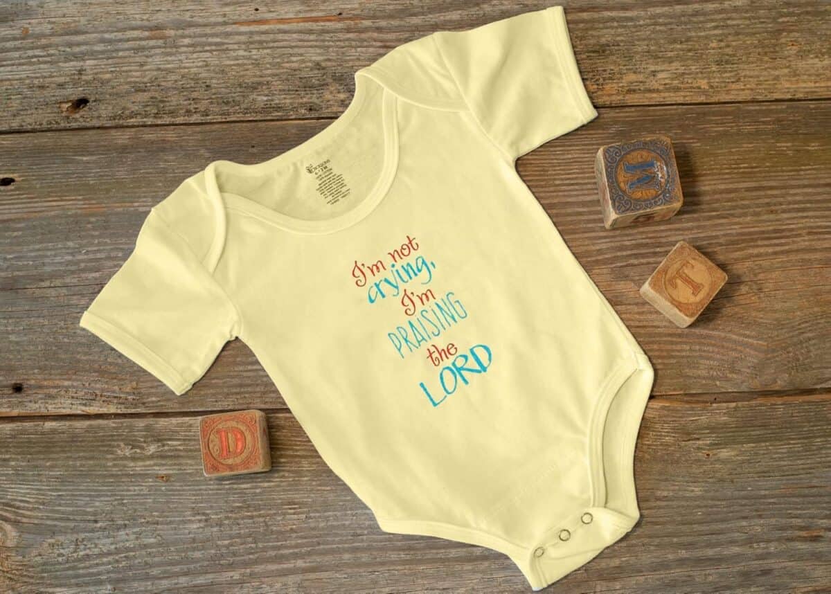 A baby's onesie displayed on a wood background with the words "I'm not crying, I'm praising the Lord" written on the shirt. Used to illustrate the post What does a first-time grandma need?