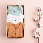 Photo illustrating some of the items a new grandparent would want to have on hand at their house. Used for the Post: What does a first-time grandma need? The photo shows a box with 3 baby outfits folded and placed inside. There is a single branch from a cotton plant to the right of the box.