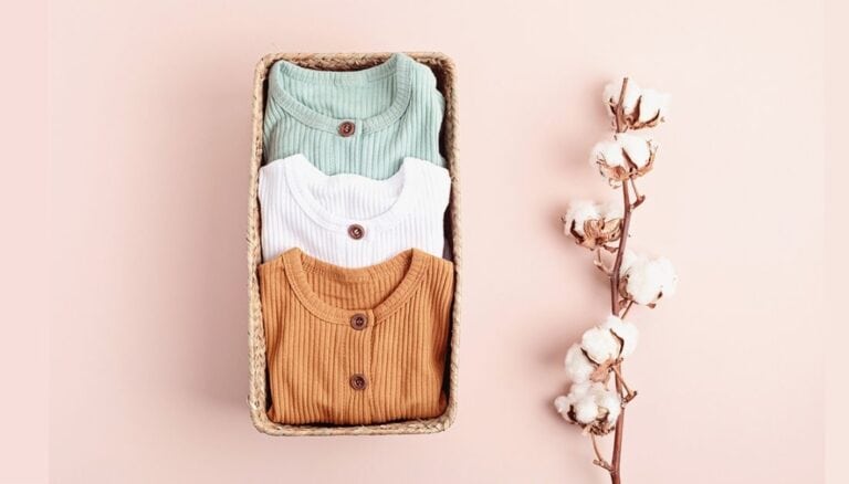 Photo illustrating some of the items a new grandparent would want to have on hand at their house. Used for the Post: What does a first-time grandma need? The photo shows a box with 3 baby outfits folded and placed inside. There is a single branch from a cotton plant to the right of the box.