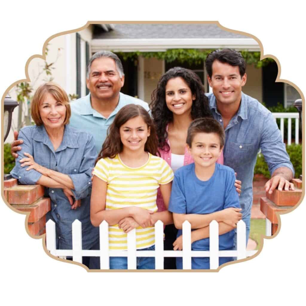 Photograph of a family with grandparents, parents, and 2 school-age children. They are outside standing in front of a house and behind a white picket fence. Used to illustrate the post How to pray for your grandchildren's faith.