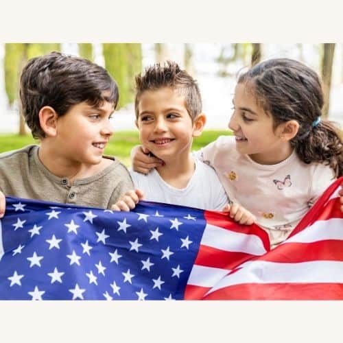 Three elementary school-aged children sitting in a park holding a flag of the USA. There are 2 boys and a girl, and they are looking at each other, smiling. Used as an in-post picture for the National Day of Prayer blog post.
