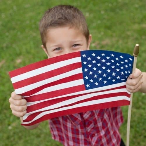 Picture of a boy of about 7 years old showing the camera his USA flag. He is holding the flag out in front of him, so his part is partially hidden. He seems happy. He is standing on grass and wearing a red checkered shirt. Used as an in-post picture for the National Day of Prayer blog post.