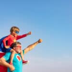 A side view of a grandfather with his grandson, who is sitting on his shoulders. The grandson is dressed in a Superhero costume that has a mask , a cape, and a red shirt with a gold star on it.. The grandfather has a mask and a red cape. They are both holding up one arm in a superhero pose. The background is a bright blue sky. Used to illustrate the blog post about grandparent's importance in their grandchildren's lives.