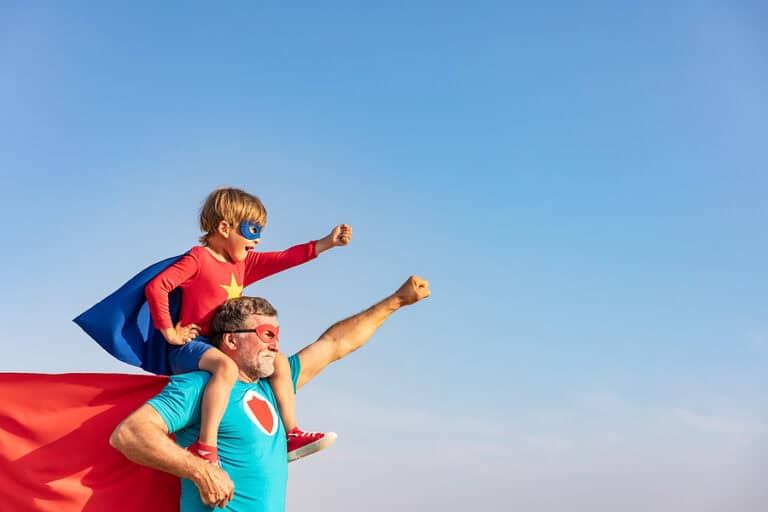 A side view of a grandfather with his grandson, who is sitting on his shoulders. The grandson is dressed in a Superhero costume that has a mask , a cape, and a red shirt with a gold star on it.. The grandfather has a mask and a red cape. They are both holding up one arm in a superhero pose. The background is a bright blue sky. Used to illustrate the blog post about grandparent's importance in their grandchildren's lives.