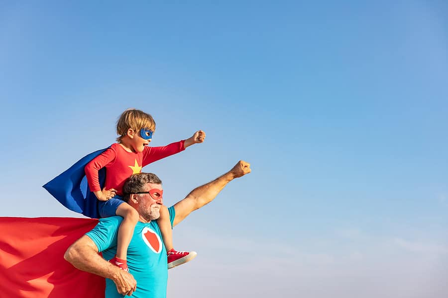 A side view of a grandfather with his grandson, who is sitting on his shoulders. The grandson is dressed in a Superhero costume that has a mask , a cape, and a red shirt with a gold star on it.. The grandfather has a mask and a red cape. They are both holding up one arm in a superhero pose. The background is a bright blue sky. Used to illustrate the blog post about grandparent's importance in their grandchildren's lives.