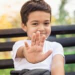 Photo of a boy aged 5-6 who is smiling shyly and holding his hand up to indicate Stop. He has brown hair and dark eyes and is wearing a t-shirt. He is sitting on a bench with greeen trees in the background. Used to illustrate the post on praying for a shy child.