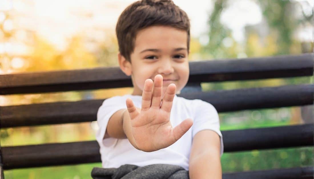 Photo of a boy aged 5-6 who is smiling shyly and holding his hand up to indicate Stop. He has brown hair and dark eyes and is wearing a t-shirt. He is sitting on a bench with greeen trees in the background. Used to illustrate the post on praying for a shy child.
