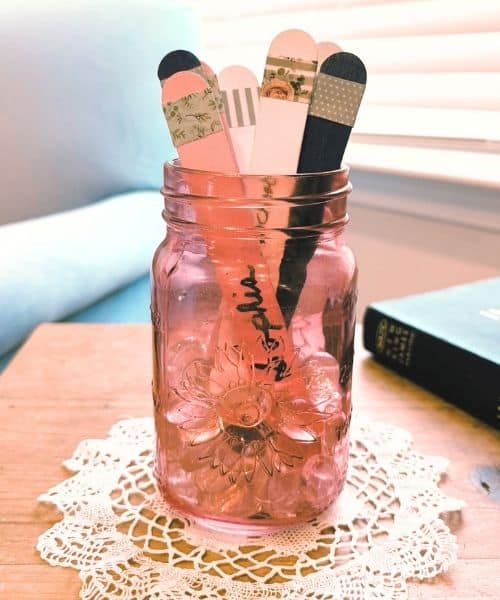 Photo of a prayer jar. It is a pink Mason jar with prayer sticks in it. The jar is sitting on a doily on a wooden table. In the background is a Bible and a blue chair.