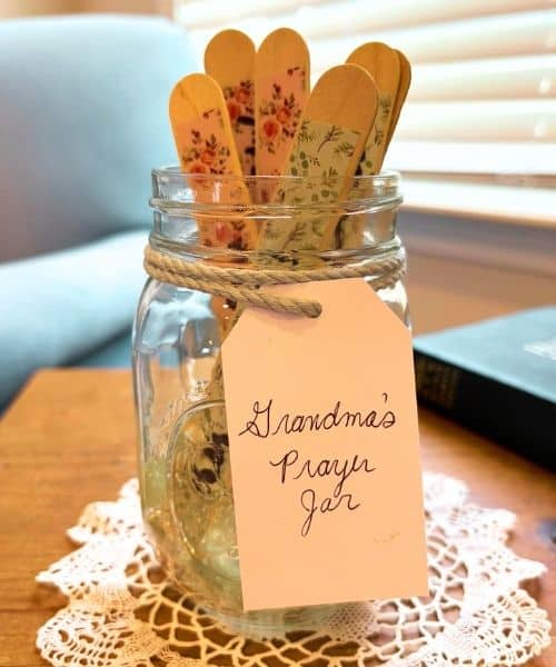 Photo of a prayer jar. The jar is a clear, glass Mason jar. It has a tag ties to it that says, "Grandma's Prayer Jar". The jar contains prayer sticks. The jar is sitting on a doily, on a wooden table. In the background is a Bible and a blue chair.