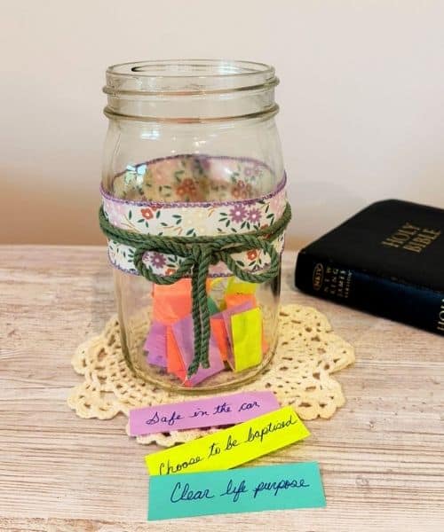 Photo of a prayer jar that uses slips of paper to record prayer requests. The jar is a clear glass Mason jar decorated with a floral ribbon and a jute string tied in a bow. The jar has colorful pieces of paper inside that contain prayer requests. There are 3 pieces of paper laying in front of the jar demonstrating what you could write on a prayer request.