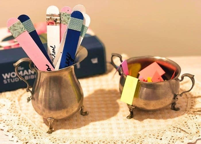 Photo of 2 prayer jars. They are a pewter cream and sugar set. The creamer contains prayer sticks and the sugar bowl has slips of paper. They are resting on a doily and there is a Bible in the background.
