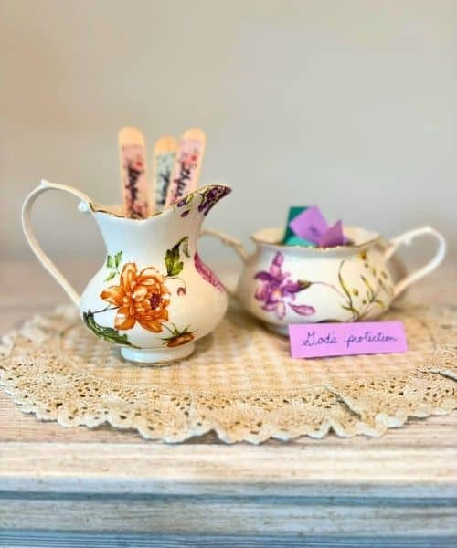 Photo of an example of 2 prayer jars. They are a cream and sugar set from a fine china set. The cream pitcher holds prayer sticks and the sugar bowls hold prayers written on paper. The jars are sitting on a doily that rests on a light wood table.