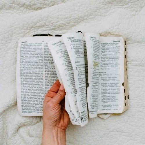 Photo of a woman's hand flipping through a Bible. The background is a white quilt. The Bible is centered, and the hand is grasping 3 pages while other pages fall back into the book. Used to illustrate the blog post 24 Bible Verses About Grandparents.
