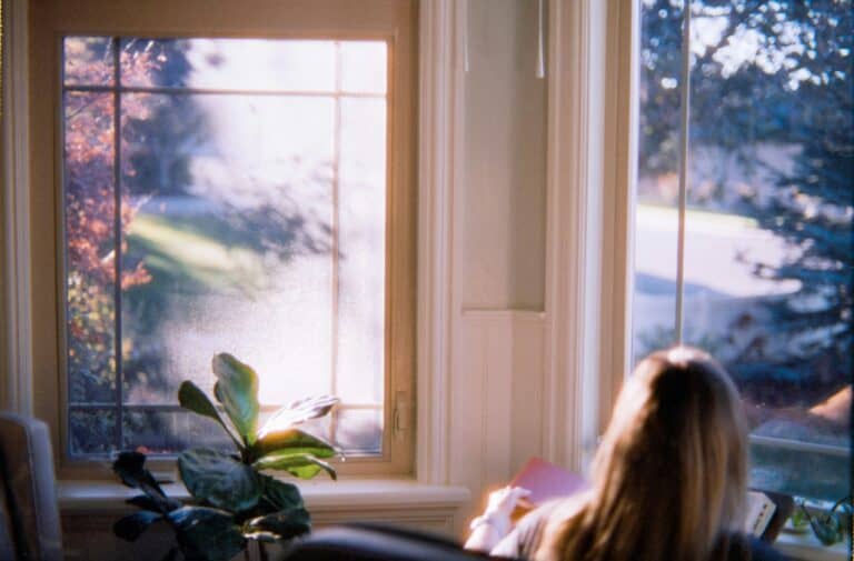 Photo of a mature woman reading her Bible. She is seated on a couch in the lower right side of the photo. In front of her is a large picture window with a view of trees and open space. On the left side is a leafy plant. Used to illustrate the post: 24 Bible verses About Grandparents.