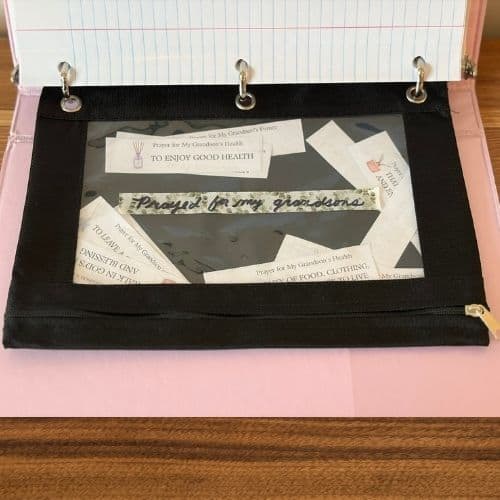 Photo of a black canvas pencil pouch. It is in a 3-ring binder. It is labeled "Prayed for my grandsons," and there are prayer slips visible through the plastic front. Used to illustrate where to keep used prayer slips for the post Prayer for a Grandson.