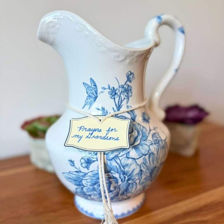 Photo of an old-fashioned ceramic pitcher. It is white with blue roses on the front. The pitcher is sitting on a wood table, and there are 2 small plants in the background. There is a cord tied around it with a label that says Prayers for my Grandsons. Used to give an example of a prayer jar for the post Prayer for a Grandson.