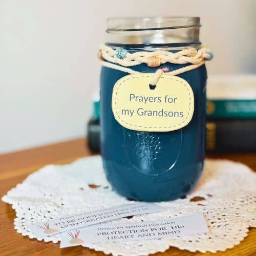 Photo of a prayer jar for the blog post Prayer for a Grandson. It is a mason jar that is painted blue. There is a braided cord around the top of the jar, and it has beads woven into it. The jar has an oval-shaped tag that says, "Prayers for my Grandsons". The jar is resting on a doily, and 2 prayer slips are sitting in front of the jar.
