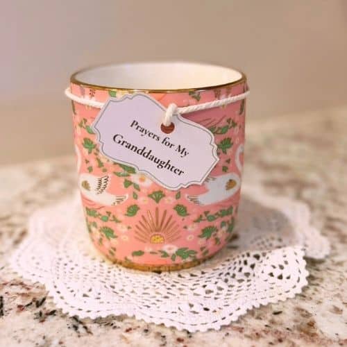 Photo of a large pink mug with swans and flowers on it. The mug is sitting on a doily which is sitting on a countertop. There is a label tied to the top of the mug with string. The label says "Prayers for My Granddaughter," Used to illustrate the post Prayer for a Granddaughter.