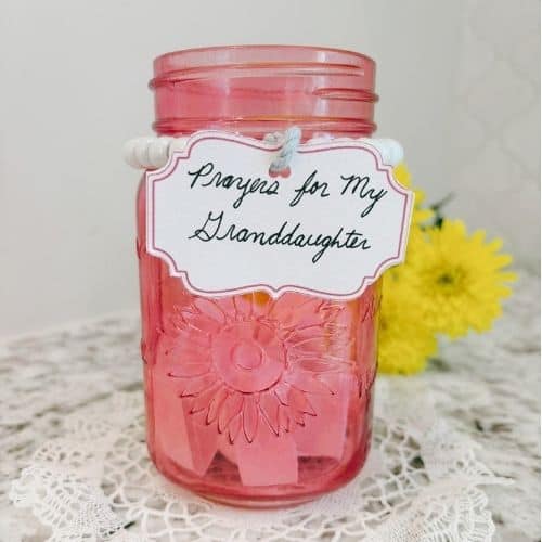 Photo of a pink Mason jar with no lid. The jar is sitting on a lacy doily on a granite countertop. In the background are some carnations. There is a beaded band around the top of the jar with a label tied to the band. The label says Prayers for My Granddaughter in cursive handwriting. Used to illustrate the post Prayer for a Granddaughter.